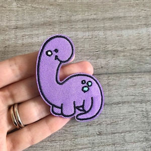 May include: Purple felt dinosaur patch with black stitching and a small blue and white design on the tail.