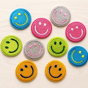 Op de afbeelding: Een collectie van tien ronde geborduurde patches met diverse smiley-gezichtjes. De patches zijn uitgevoerd in felle kleuren zoals blauw, roze, limoengroen, grijs en geel, elk met een geborduurde lachende uitdrukking in een contrasterende draadkleur.