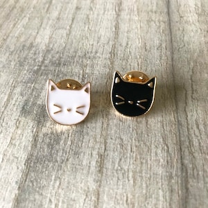 May include: Two cat-shaped enamel pins. One pin is white with a black nose and whiskers, the other is black with a white nose and whiskers. Both pins have gold-colored metal trim and backing, and are set against a gray wood grain surface.