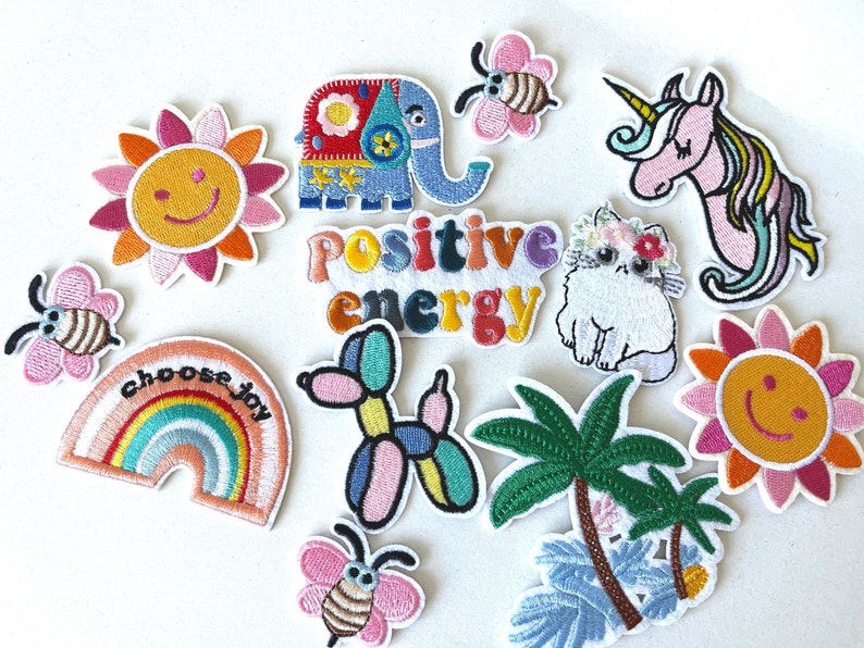 May include: A collection of embroidered patches featuring a smiling sun, a blue elephant, a bee, a unicorn, a cat, a rainbow, a balloon dog, palm trees, and the words "positive energy" and "choose joy". The patches are colorful and whimsical.