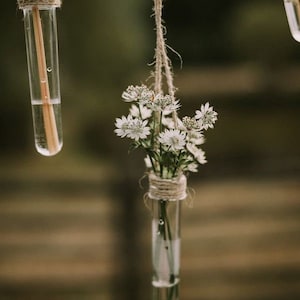 May include: White flowers in a glass test tube hanging from a string. The test tube is tied with twine and the flowers are arranged in a bouquet.