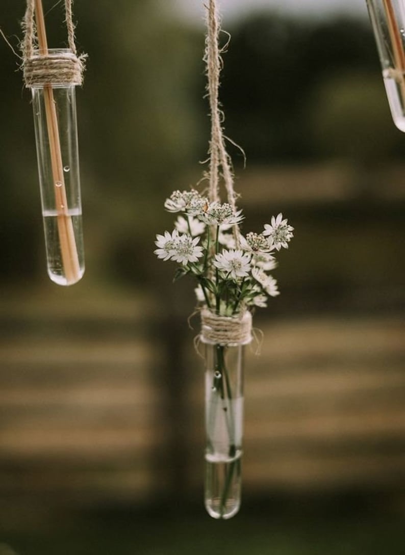 May include: White flowers in a glass test tube hanging from a string. The test tube is tied with twine and the flowers are arranged in a bouquet.