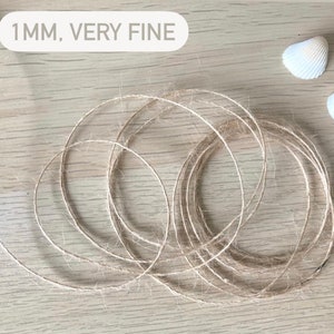May include: A close-up of a natural fiber twine, 1 mm thick, labeled "1MM, VERY FINE". The twine is coiled on a light wood surface. Two white seashells are in the background.