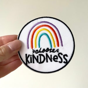 May include: A circular, white embroidered patch featuring a rainbow design and the words "choose KINDness" in black. The rainbow has purple, orange, yellow, and teal arches. The patch is edged with a black border.