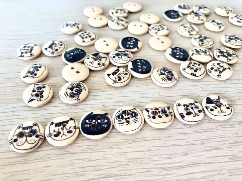 May include: A collection of wooden buttons with black and white cat faces. The buttons are various sizes and shapes, with some having two holes and others having four holes.