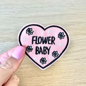 May include: Pink heart-shaped embroidered patch with black stitching and white flowers. The patch says "Flower Baby" in black lettering.