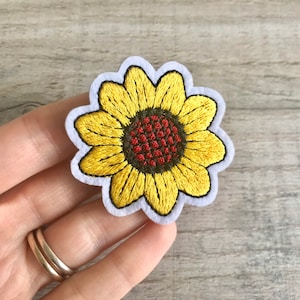 Könnte beinhalten: Ein gestickter Sonnenblumen-Patch in Gelb mit einer roten Mitte. Der Patch ist zum Aufbügeln und kann zum Dekorieren von Kleidung, Taschen oder anderen Gegenständen verwendet werden.