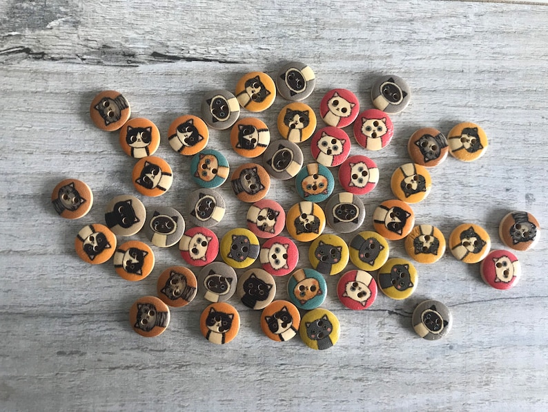 May include: A collection of colorful wooden buttons featuring various cat designs. The buttons are round and have a flat back. They are perfect for adding a whimsical touch to crafts and sewing projects.
