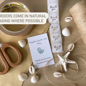 May include: A close-up of a brown paper package with a roll of brown packing tape, a pair of wooden handled scissors, and a small white card with the text "Supplies by the Sea. Etsy" and "Thank you for your lovely purchase!"  The package is decorated with seashells and a white starfish. There are also five white round stickers with gold lettering that say "Thank you".