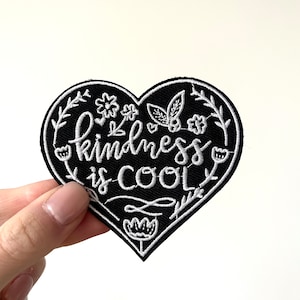 May include: Black heart-shaped embroidered patch with white stitching. The patch features the text "kindness is cool" surrounded by small white flowers and leaves.
