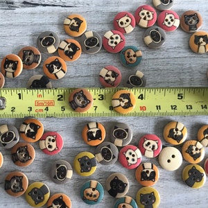 May include: A collection of wooden buttons with various cat designs. The buttons are painted in a variety of colors, including black, white, brown, orange, and pink. The buttons are arranged on a wooden surface with a measuring tape.