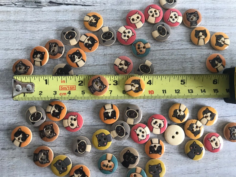 May include: A collection of wooden buttons with various cat designs. The buttons are painted in a variety of colors, including black, white, brown, orange, and pink. The buttons are arranged on a wooden surface with a measuring tape.