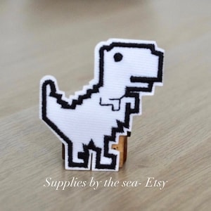 以下が含まれることがあります： Google Chrome のオフラインゲームのクラシックなピクセル化された恐竜の白い刺繍パッチ。パッチは木製の背景の上にあります。