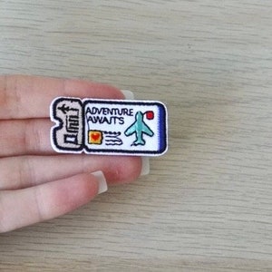 May include: Embroidered patch in the shape of a ticket with the words "Adventure Awaits" and a small airplane. The patch is white with blue, red, and black details. The patch is held in a hand.