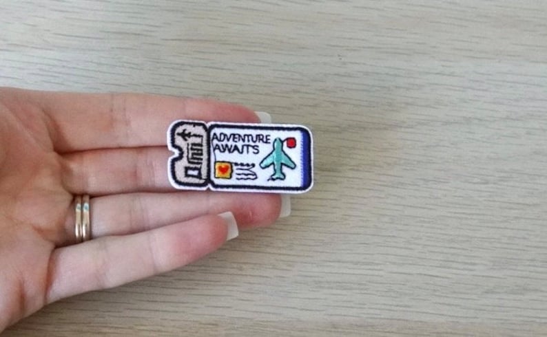 May include: Embroidered patch in the shape of a ticket with the words "Adventure Awaits" and a small airplane. The patch is white with blue, red, and black details. The patch is held in a hand.