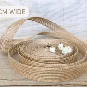 May include: A roll of natural jute ribbon, 2 cm wide, with a textured woven pattern. The ribbon is coiled and has a few small white flowers on top.