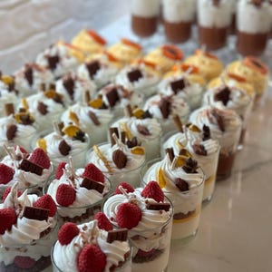 Op de afbeelding: Een verzameling gelaagde desserts in heldere glazen. De desserts zijn voorzien van witte slagroom, chocoladeschilfers en vers fruit, waaronder aardbeien en sinaasappelschijfjes. Sommige hebben een chocoladebasis, andere een gele basis.