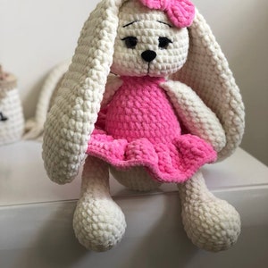 Könnte beinhalten: Ein gehäkeltes Hasenspielzeug, das ein rosa Kleid und eine rosa Schleife trägt. Der Hase hat weißes Fell und schwarze Augen.