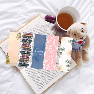 Puede incluir: Cuatro marcapáginas diferentes con varios diseños. Un marcapáginas presenta una pila de libros con el texto "Just one more chapter". Otro marcapáginas tiene un fondo azul con estrellas blancas y el texto "Reading is dreaming with open eyes". El tercer marcapáginas tiene un fondo rosa con diseños florales blancos. El último marcapáginas tiene un fondo blanco con diseños florales verdes y amarillos.