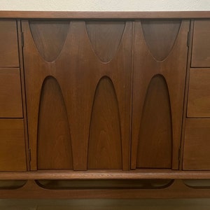 May include: A mid-century modern dresser with a wood veneer finish and a unique carved design on the doors. The dresser has four drawers on each side and a lower shelf. The dresser is made of solid wood and has a rich brown color.
