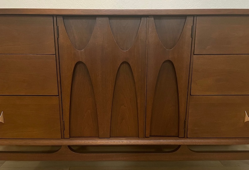 May include: A mid-century modern dresser with a wood veneer finish and a unique carved design on the doors. The dresser has four drawers on each side and a lower shelf. The dresser is made of solid wood and has a rich brown color.