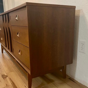 May include: A mid-century modern dresser with six drawers and brass hardware. The dresser is made of wood and has a warm brown finish. It is sitting on a wooden floor.