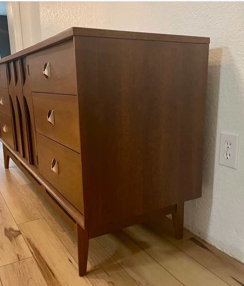 May include: A mid-century modern dresser with six drawers and brass hardware. The dresser is made of wood and has a warm brown finish. It is sitting on a wooden floor.