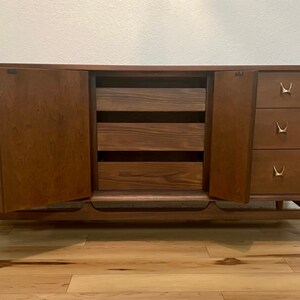 May include: A mid-century modern dresser with six drawers and two cabinet doors. The dresser is made of wood and has a dark brown finish. The drawers have brass handles with a unique design. The cabinet doors are open, revealing three shelves inside.
