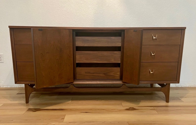 May include: A mid-century modern dresser with six drawers and two cabinet doors. The dresser is made of wood and has a dark brown finish. The drawers have brass handles with a unique design. The cabinet doors are open, revealing three shelves inside.