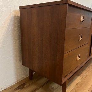 May include: A mid-century modern dresser with three drawers and a wood grain finish. The dresser has brass hardware with a unique triangular shape.