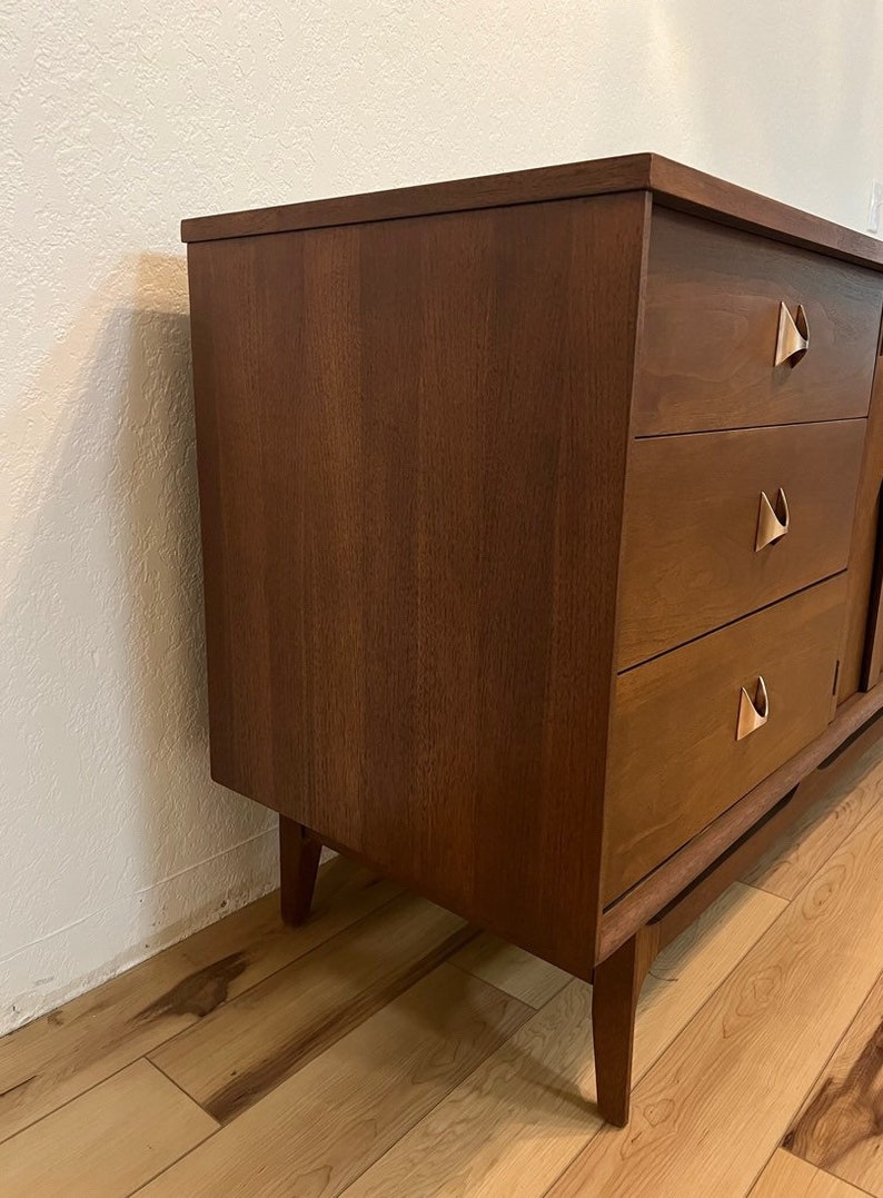 May include: A mid-century modern dresser with three drawers and a wood grain finish. The dresser has brass hardware with a unique triangular shape.