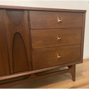 May include: A mid-century modern dresser with three drawers and a cabinet door. The dresser is made of dark wood with brass hardware. The dresser has a unique design with curved edges and a slightly angled base.