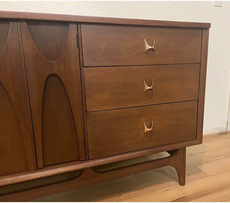 May include: A mid-century modern dresser with three drawers and a cabinet door. The dresser is made of dark wood with brass hardware. The dresser has a unique design with curved edges and a slightly angled base.