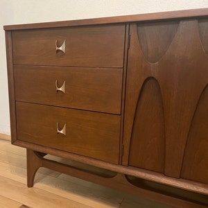 May include: A mid-century modern dresser with three drawers and a cabinet door. The dresser is made of wood and has a dark brown finish. The drawers have silver metal handles in the shape of a stylized "H".