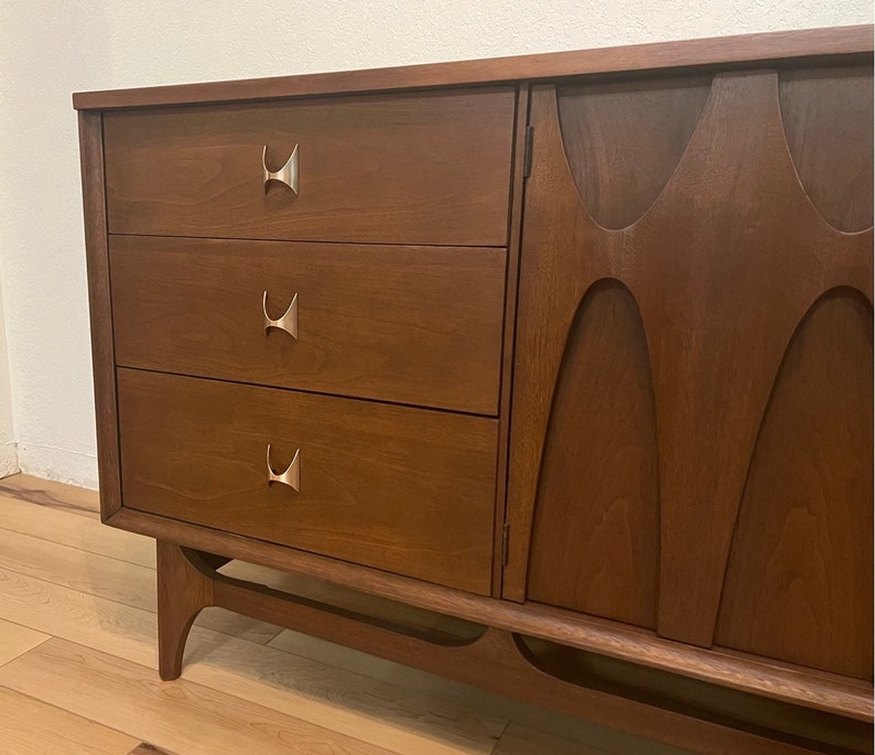 May include: A mid-century modern dresser with three drawers and a cabinet door. The dresser is made of wood and has a dark brown finish. The drawers have silver metal handles in the shape of a stylized "H".