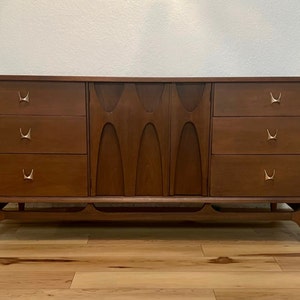 May include: A mid-century modern dresser with six drawers and a unique carved wood design on the front. The dresser is made of dark wood and has brass hardware.