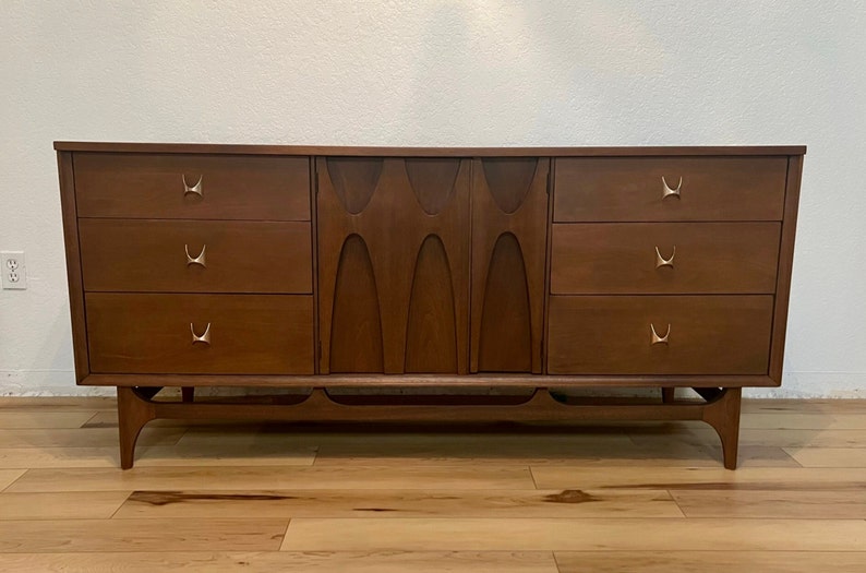 May include: A mid-century modern dresser with six drawers and a unique carved wood design on the front. The dresser is made of dark wood and has brass hardware.
