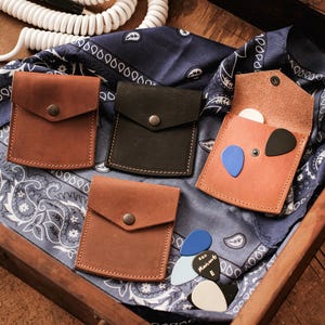 Könnte beinhalten: Vier Gitarrenplektrumhalter aus braunem und schwarzem Leder, mit einem blauen Paisley-Bandana und Gitarrenplektren. Ein Halter ist geöffnet und zeigt Plektren in verschiedenen Farben. Ein weißes Spiralkabel befindet sich im Hintergrund.