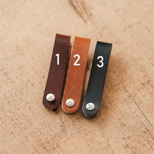 May include: Three leather key fobs in brown, tan, and black, each with a silver rivet and a number. The fobs are arranged diagonally on a wooden surface. The numbers 1, 2, and 3 are printed on the fobs.