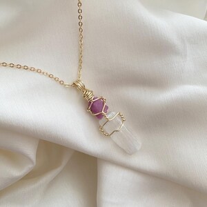 May include: A gold chain necklace with a pendant. The pendant has a pink bead, a clear bead, and a white crystal, all wrapped in gold wire. The necklace is displayed on white, silky fabric.