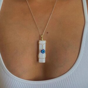 Pode incluir: Um pingente retangular de cristal de selenita branco envolto em fio dourado, apresentando uma pequena conta de olho grego azul e branco fixada na frente. O pingente pende de uma corrente fina dourada, criando um colar detalhado e em camadas.