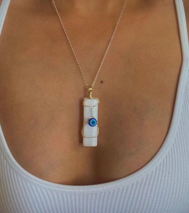Pode incluir: Um pingente retangular de cristal de selenita branco envolto em fio dourado, apresentando uma pequena conta de olho grego azul e branco fixada na frente. O pingente pende de uma corrente fina dourada, criando um colar detalhado e em camadas.