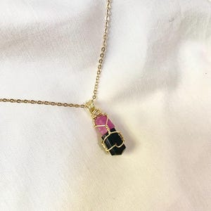May include: A gold chain necklace with a pendant. The pendant has a pink and black crystal encased in gold wire. The necklace is displayed on a white background. The pendant is rectangular.