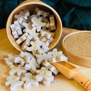 Peut inclure: Un récipient rond en carton rempli de fondants de cire gris clair en forme de bonshommes en pain d'épices, saupoudrés de paillettes dorées. Une pelle en bois repose parmi les figurines, éparpillées à côté du couvercle du récipient.