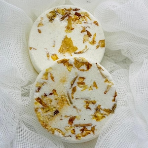 May include: Two round, white bath bombs with a textured surface, adorned with dried yellow flower petals. The bath bombs rest on a white, gauzy fabric, creating a soft, spa-like aesthetic. The overall theme is self-care and relaxation.