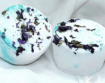 May include: Two white bath bombs with a light blue hue and decorated with dried purple flowers and green herbs. The bath bombs are round and sit on a white fabric background. The bath bombs are a self-care product.