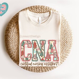 May include: A white t-shirt featuring a Christmas-themed design that reads "CNA certified nursing assistant". The letters are decorated with candy canes, baubles, and Christmas trees.