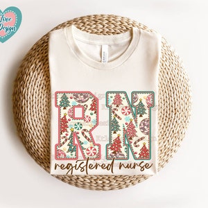 May include: A white t-shirt with a Christmas-themed design featuring the letters "RN" with a candy cane and Christmas tree pattern. The text "registered nurse" is below the letters.