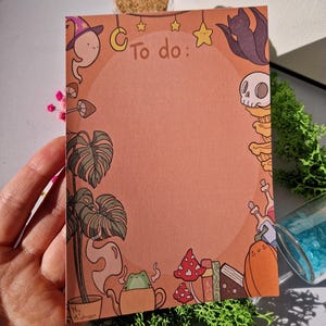 Może przedstawiać: Brzoskwiniowy notatnik z napisem "To do:" na górze. Notatnik jest ozdobiony fantazyjnymi ilustracjami ducha, nietoperza, czaszki, grzybów i żaby w czajniczku. Notatnik jest trzymany w dłoni.