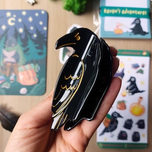 K&ouml;nnte beinhalten: Eine schwarze Haarspange in Form eines Raben mit goldfarbenen Linien, die den Fl&uuml;gel und den Schnabel des Vogels nachzeichnen. Die Spange hat eine gl&auml;nzende Oberfl&auml;che und einen stabilen Klappmechanismus, um das Haar sicher mit den Zinken zu halten.